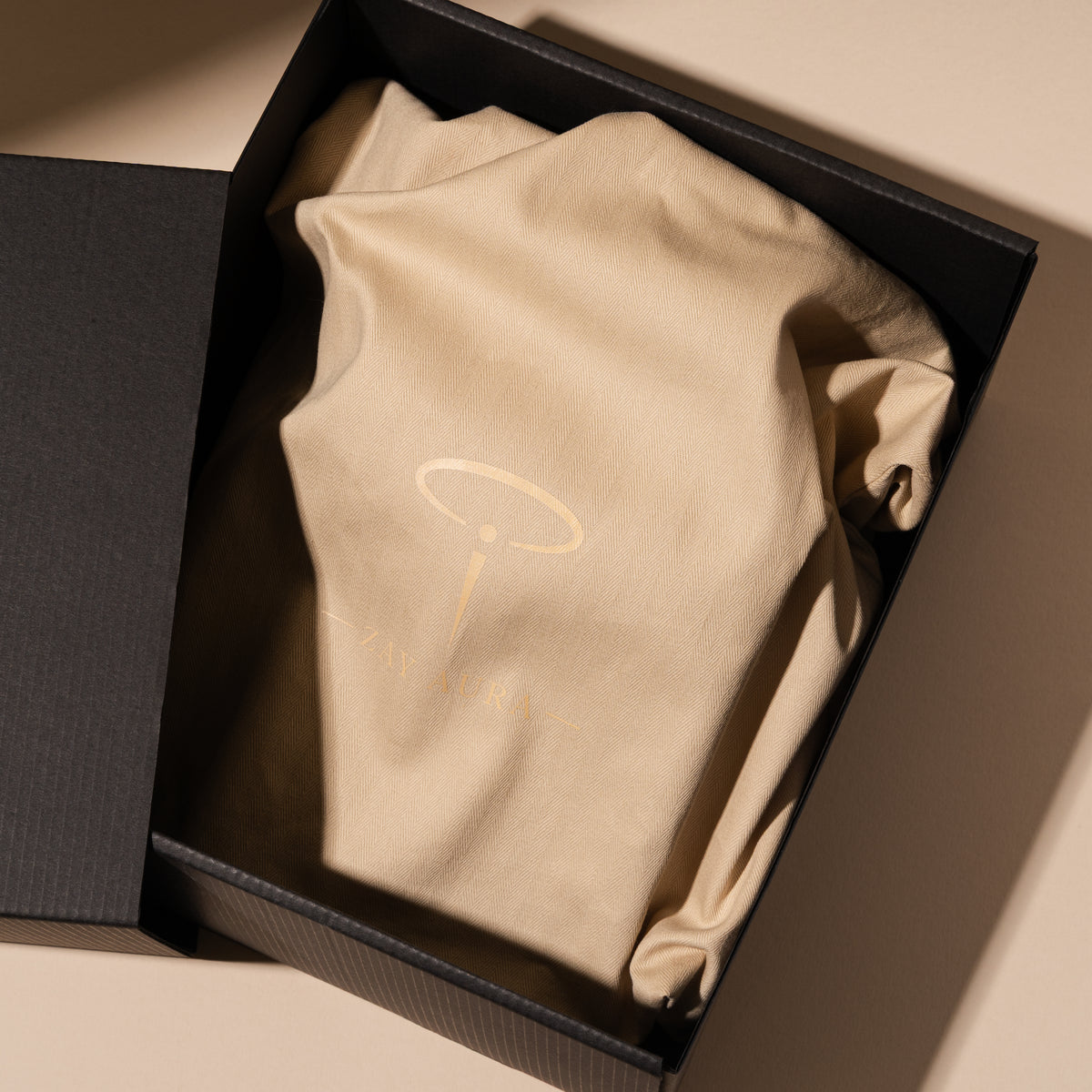 Beige ZAY AURA fabric dust bag with a gold halo logo inside an open black presentation box, photographed from above.